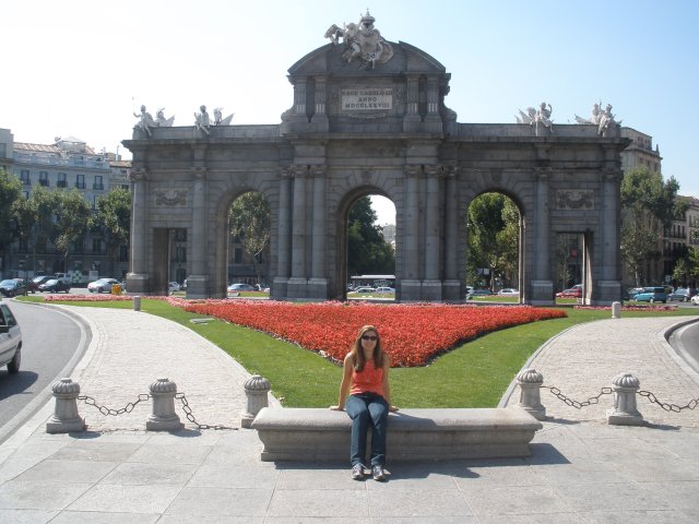 Estou sentada em uma praça e ao fundo a Porta de Alcalá, possui duas portas retangulares que estão ao lado de três arcos